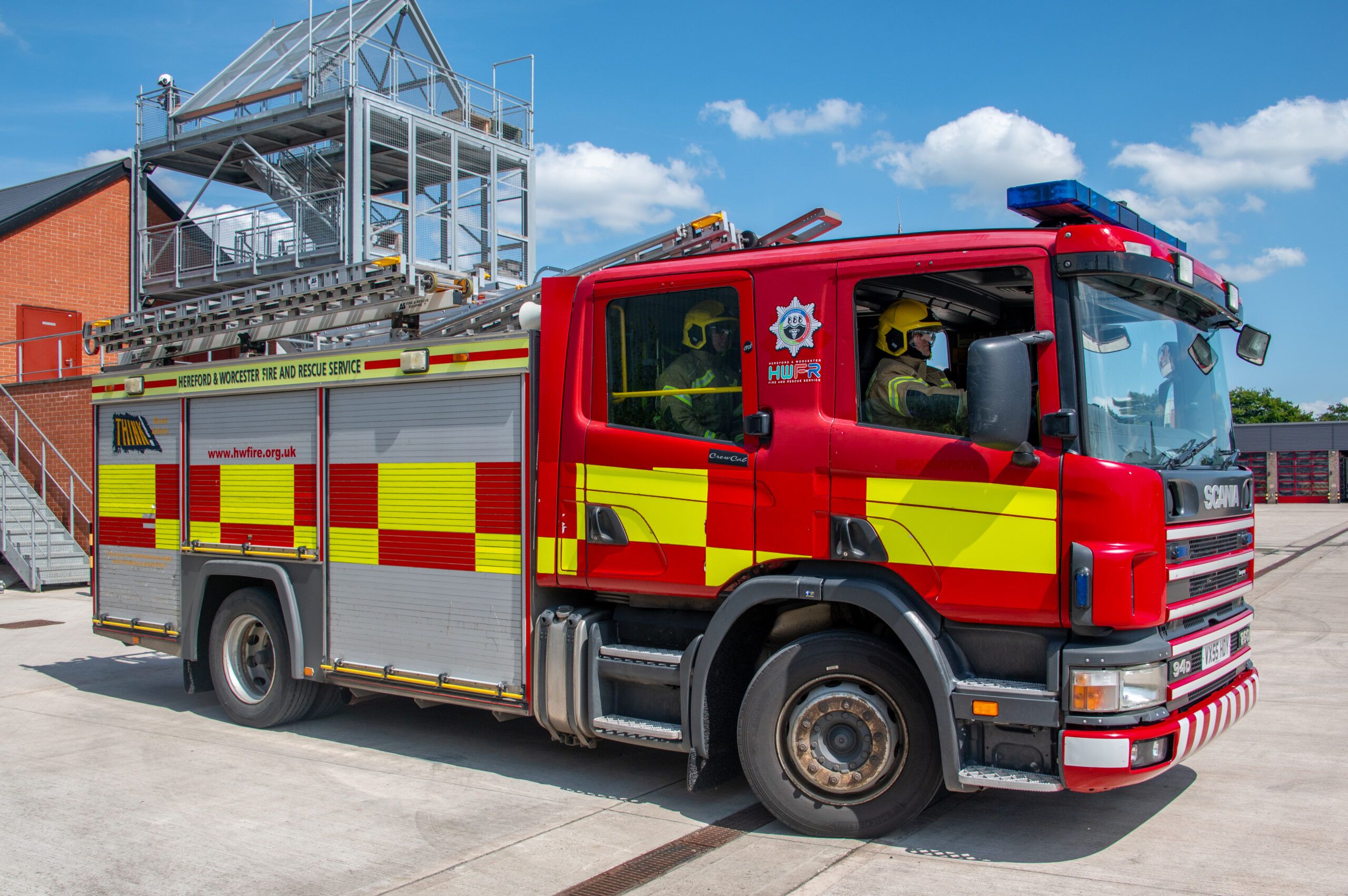 A fire engine being driven by a firefighter wearing PPE and a helmet. A firefighter in PPE is visible through the rear window of the cab.